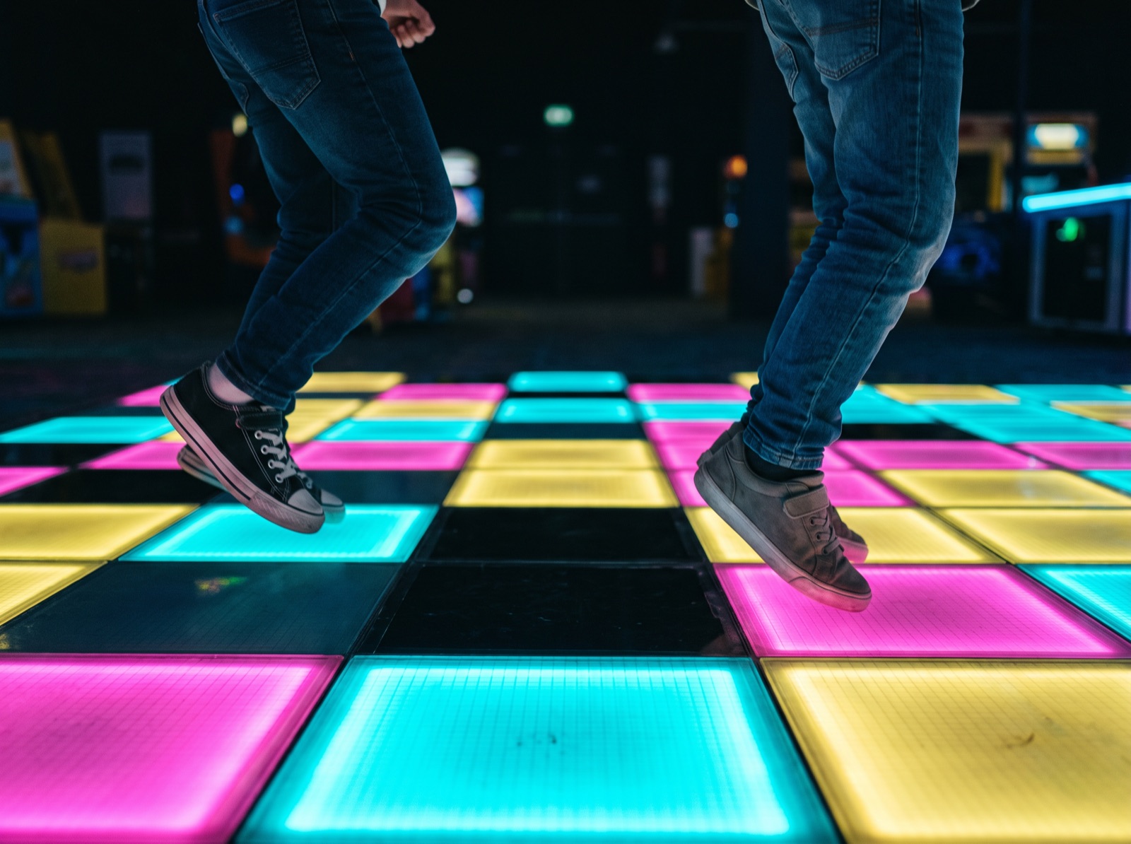 Two children mid-jump over a floor of glowing pink, cyan and yellow tiles, seen from the knees down