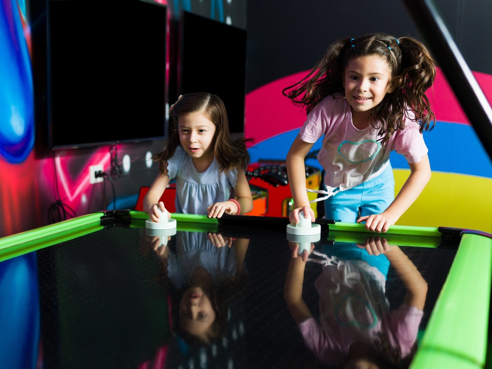 Two girls playing air hockey beside a brightly painted wall