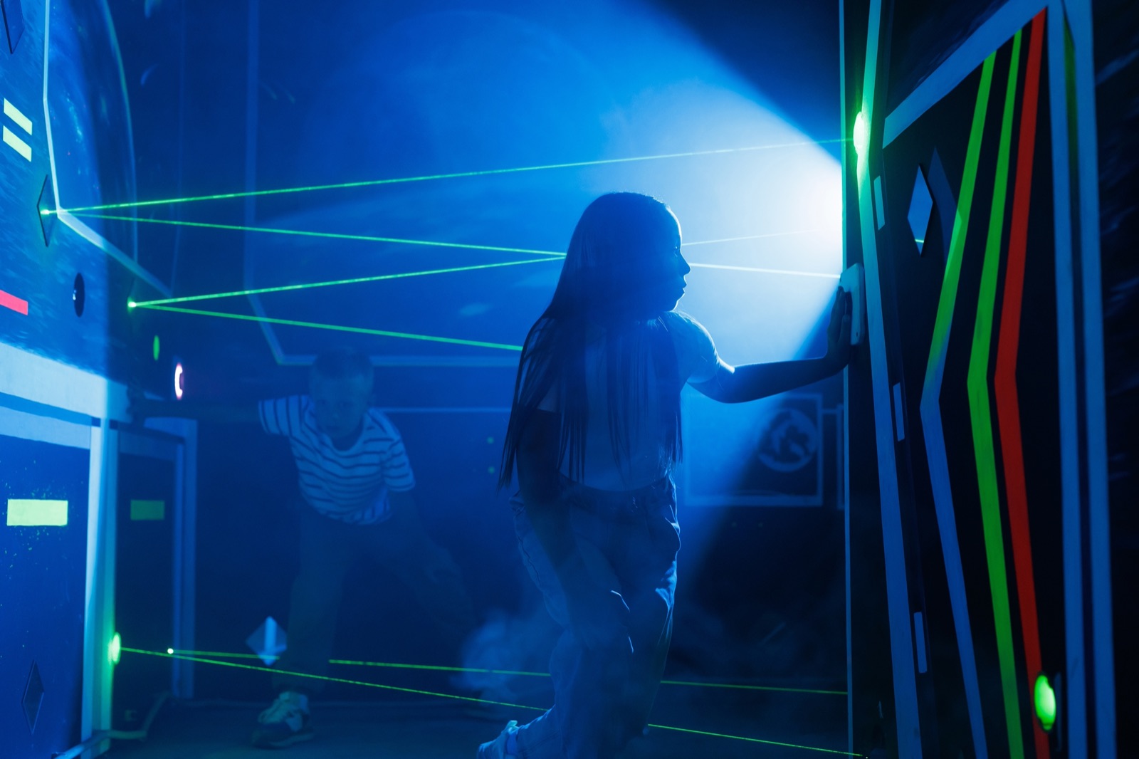 A girl stepping between green laser beams in a dark room, a boy ducking under them behind her
