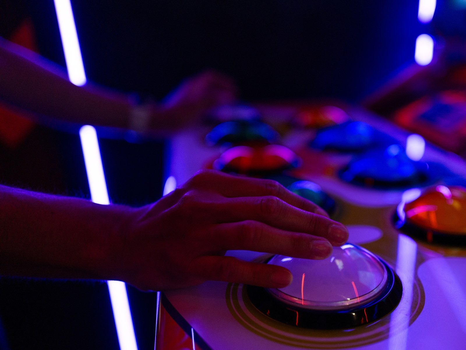 A hand pressing a glowing dome button on a game console, another player's arm beside it